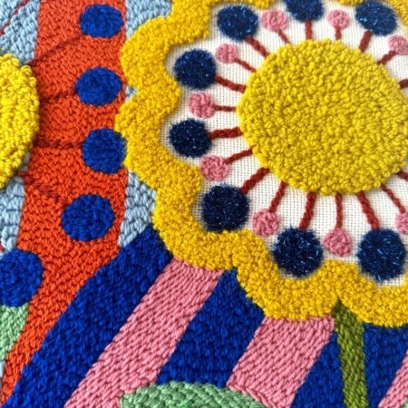 Tableau textile brodé représentant trois grandes fleurs stylisées sur un fond de motifs arlequin colorés et géométriques, réalisé en punch needle