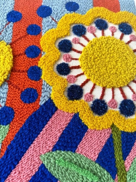 Tableau textile brodé représentant trois grandes fleurs stylisées sur un fond de motifs arlequin colorés et géométriques, réalisé en punch needle