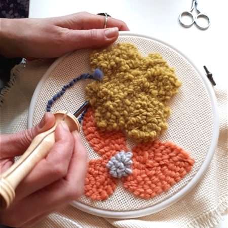 Personne utilisant une aiguille Oxford pour puncher de la laine jaune, orange et bleue sur un tambour de broderie.