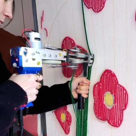 Personne utilisant un tufting gun bleu pour tracer une ligne verte sur une toile tendue décorée de fleurs roses.