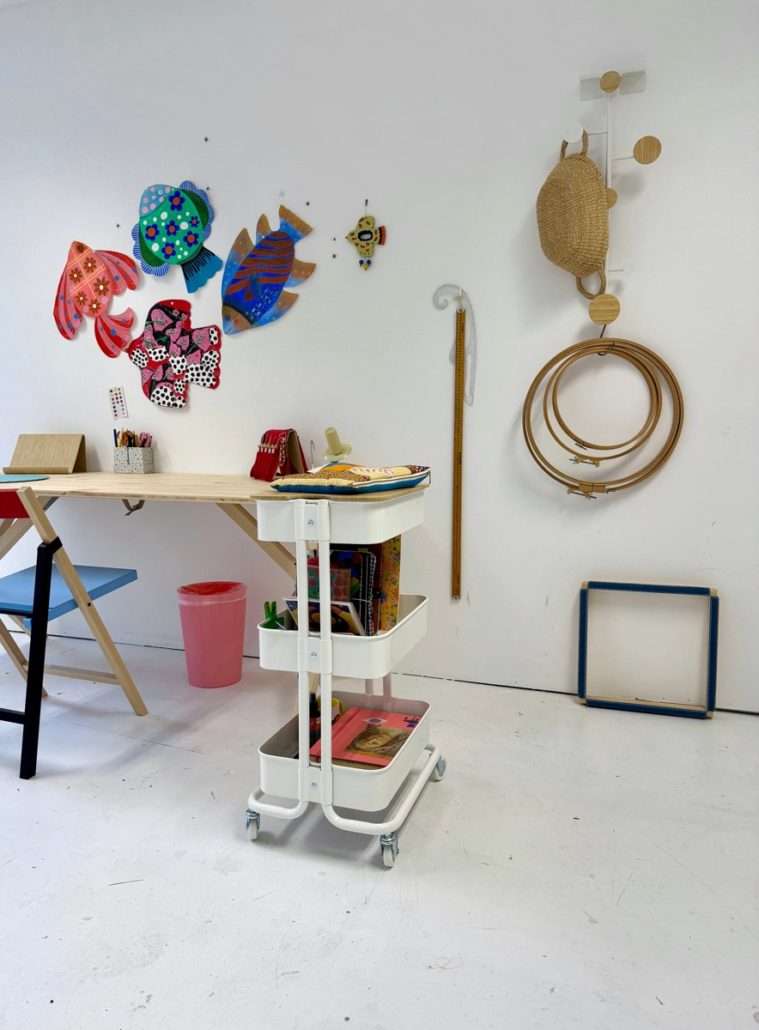 Vue du bureau 220 d'Agnès Pironon, avec des poissons textiles colorés au mur, une table de travail, un chariot de matériel et des tambours à broder suspendus.
