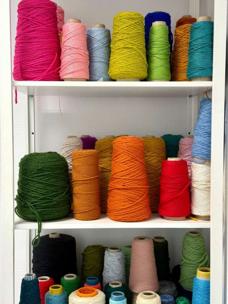 Bobines de laine rose, jaune, orange, verte, bleue et rouge disposées sur trois étagères blanches dans l'atelier textile d'Agnès Pironon.