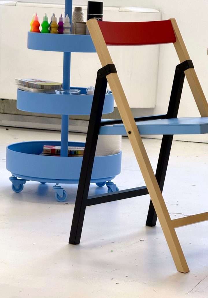 Chaise en bois à assise bleue et dossier rouge devant un chariot bleu rempli de matériel artistique, dans l'atelier d'Agnès Pironon.