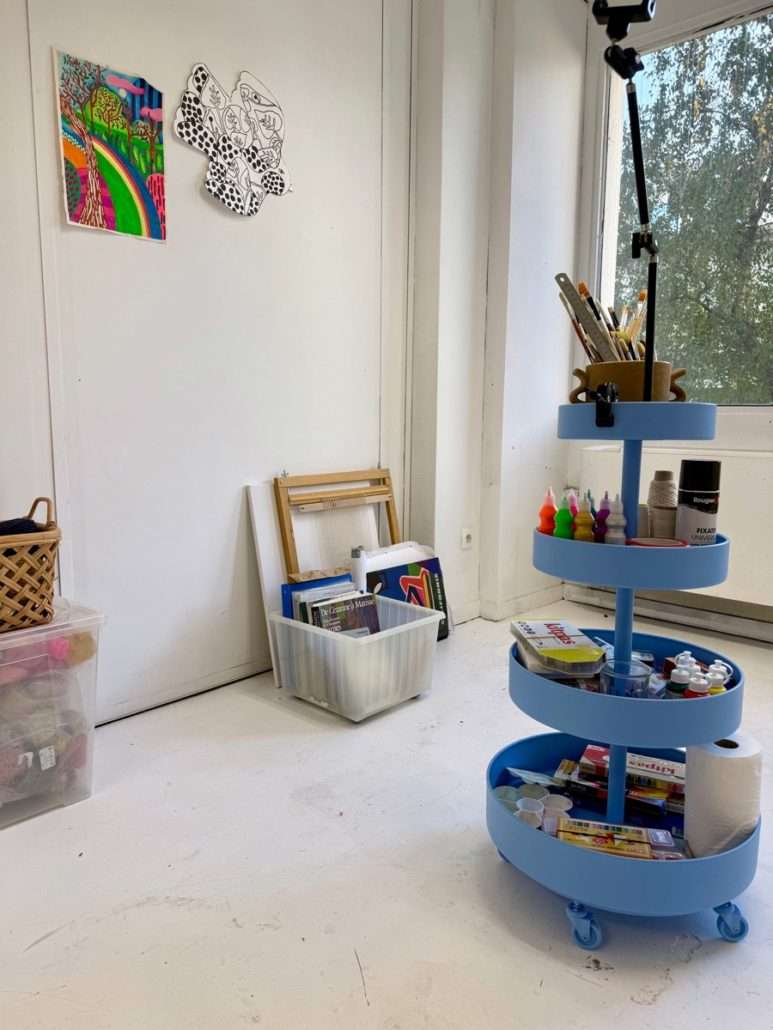 Chariot bleu contenant des pinceaux, des peintures et du matériel artistique dans le bureau 220 d'Agnès Pironon, près d'une fenêtre et de deux œuvres accrochées au mur.