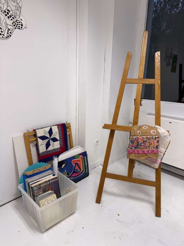 Coin de l'atelier d'Agnès Pironon avec un chevalet en bois, des textiles et une caisse de livres d'art.
