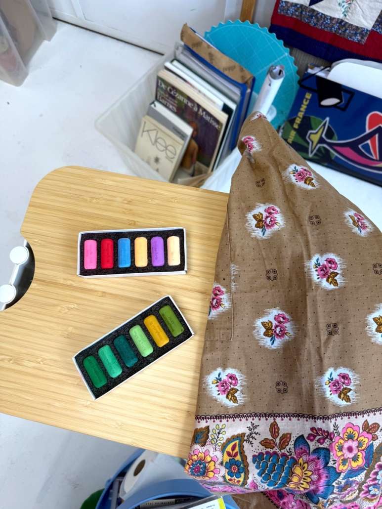 Deux boîtes de pastels aux couleurs vives posées sur une table en bois, à côté d'un tissu à motifs floraux dans l'atelier d'Agnès Pironon.