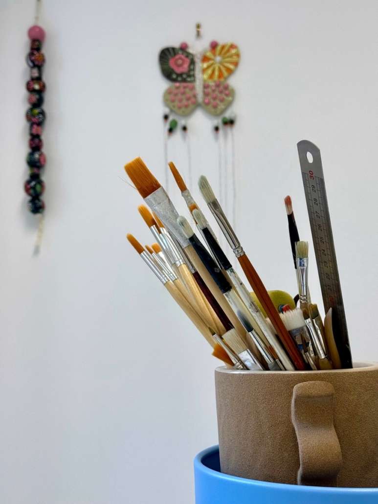 Pinceaux de peinture et règle métallique dans un pot en céramique, avec des objets textiles colorés en arrière-plan.