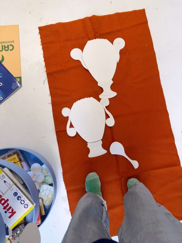 Vue plongeante sur des formes découpées dans du papier blanc, disposées sur un tissu orange au sol, avec du matériel de peinture et les pieds de l'artiste visibles.