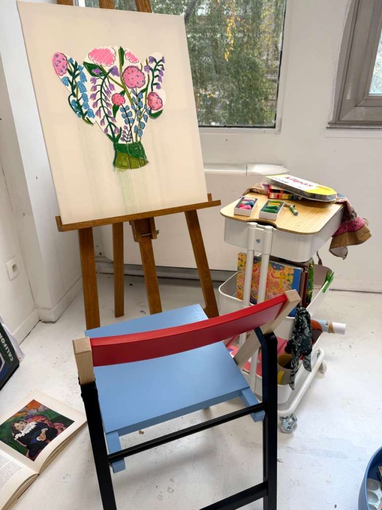 Composition florale en forme de vase sur une toile posée sur un chevalet, près d'une chaise colorée et d'un chariot de matériel artistique dans l'atelier d'Agnès Pironon.